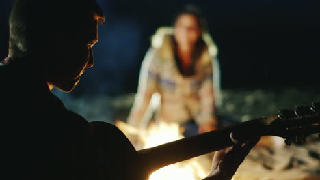 man playing guitar to his girlfriend sit around the campfire in the evening next to each other