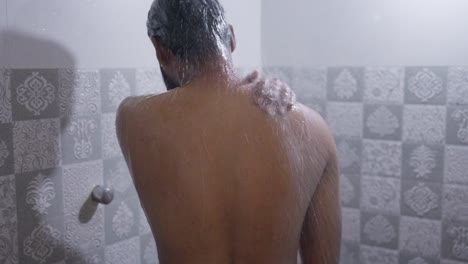 a back shot of a south asian man rubbing his body with hot water in the shower under bright natural light