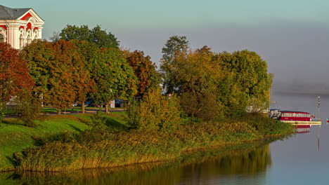 A-relaxing-landscape-near-Jelgava-Castle:-boats-anchored-near-a-pierce,-trees-and-bushes-grow-along-the-river-bank,-and-a-piece-of-the-mansion-is-seen-behind-the-trees