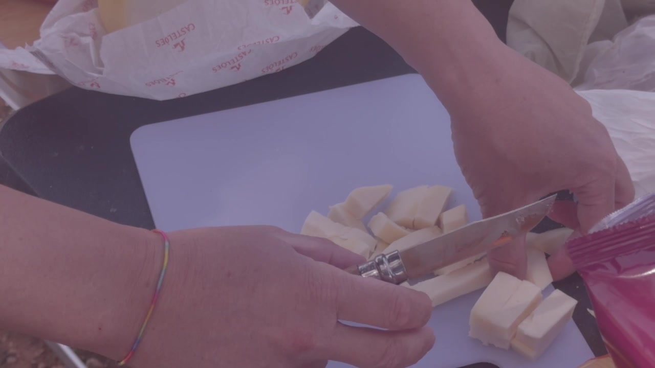 Manos cortan queso claro en pequeños cubos sobre una tabla blanca durante una comida de campamento al aire libre
