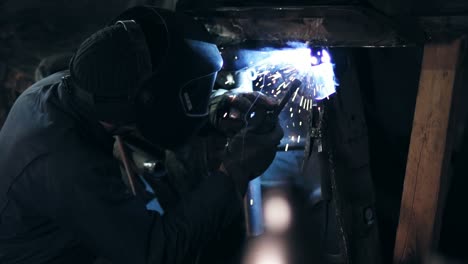 slow-motion footage of workman in helmet welding something on bottom side of an old car construction.