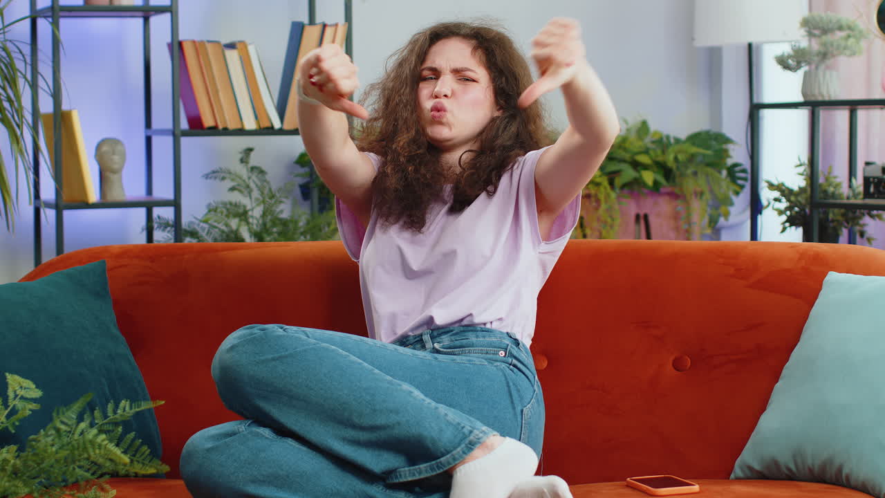 화가 난 젊은 여성은 엄지손가락을 내려놓고, 나쁜 일을 싫어하고, 승인하지 않고, 집에서 불만족스러운 피드백을합니다.