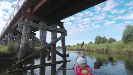 Tracking-slow-motion-shot-of-a-man-in-a-red-kayak-paddling-under-an-old-bridge-on-a-river