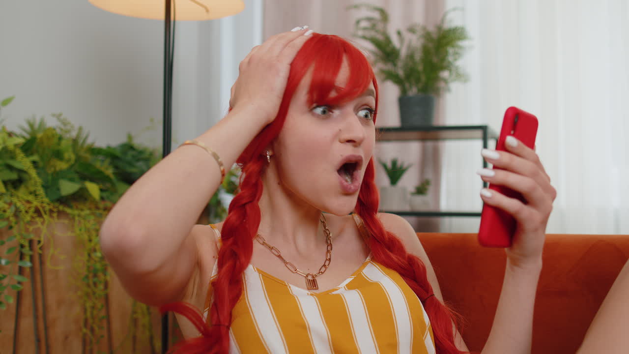 집에서 소파에 있는 어린  ⁇ 은 머리 소녀 스마트폰을 사용하고 타이핑하고 브라우징하고 와우라고 말합니다.