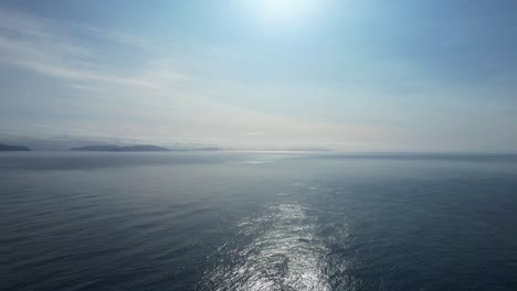 Aerial-pedestal-above-Atlantic-Ocean-with-bright-sun-beam-ray-across-water,-Faroe-Islands-in-distance