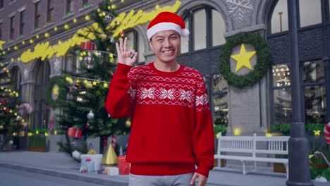 asian man in santa suit showing ok gesture and smiling while standing in christmas night