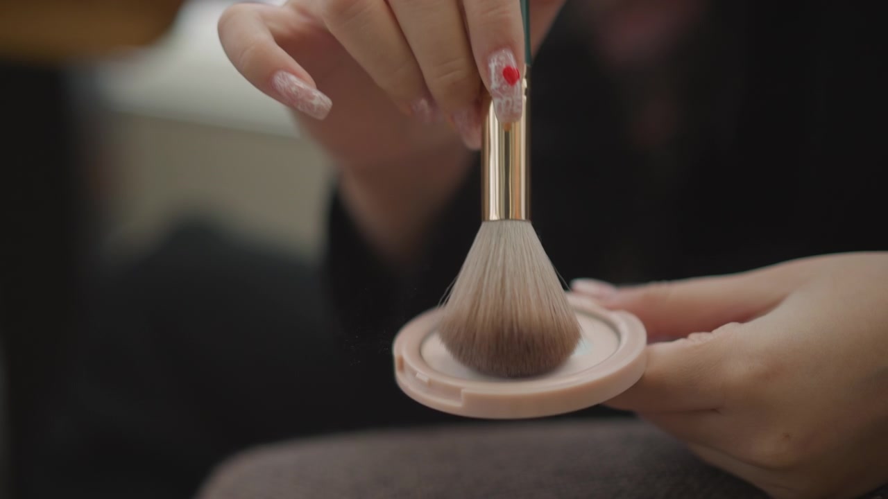 Hands press and rotate rounded powder brush inside compact, showing visible cosmetic loading, powder displacement, gentle bristle splay, painted nails with small red heart detail, and soft studio