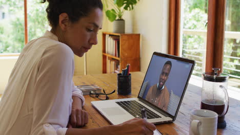 mujer de negocios biracial sentada en el escritorio usando una computadora portátil haciendo una llamada de video con un colega masculino