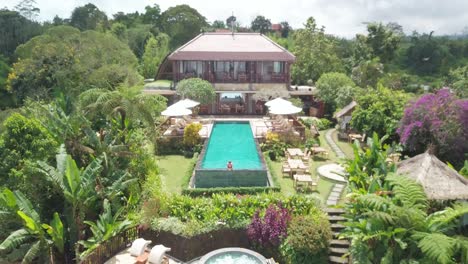 aerial view, young woman in swimming pool of luxury villa on bali island, exotic tropical vacation concept, pull back revealing drone shot