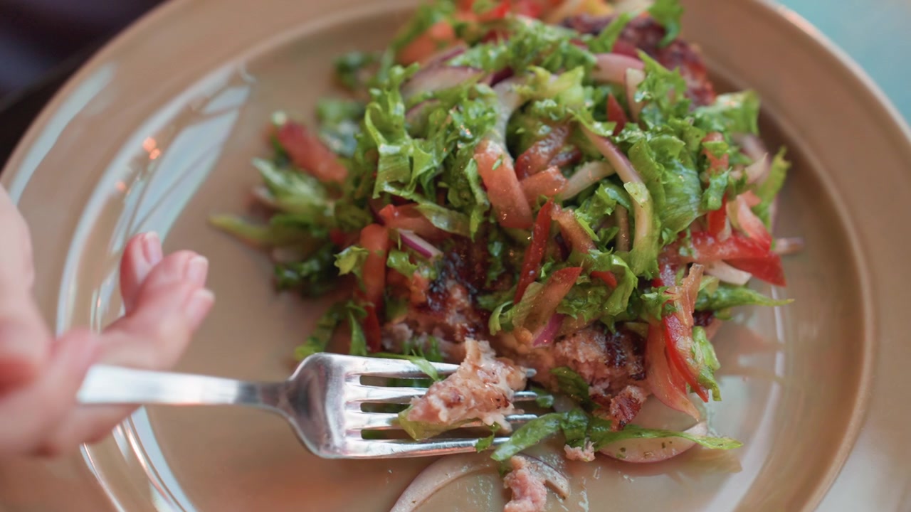 Hand holding fork repeatedly picks up and moves food from plate containing salad greens tomatoes onions meat patties or similar ingredients. close up view focuses on interaction