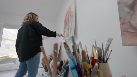 artist stands painings canvas in home studio, foregrounded by pots of brushes