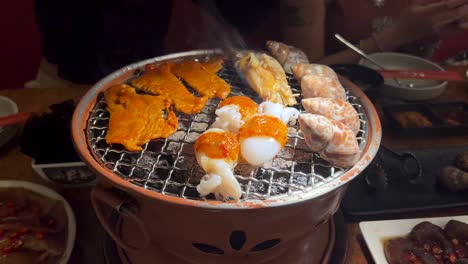 seafood and assorted meats cook on a tabletop charcoal grill in a restaurant setting, with visible smoke, warm lighting, and diners in the background