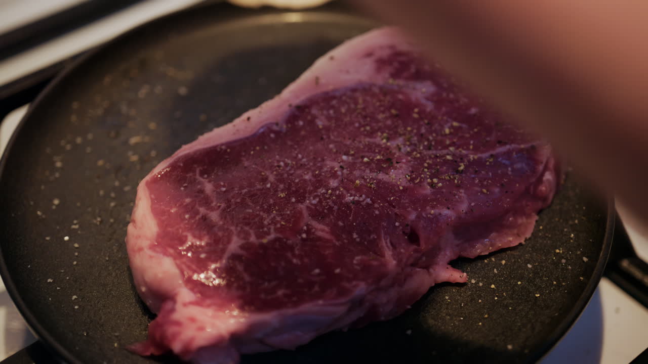 Close up of a raw beef steak being seasoned with salt and black pepper in a pan