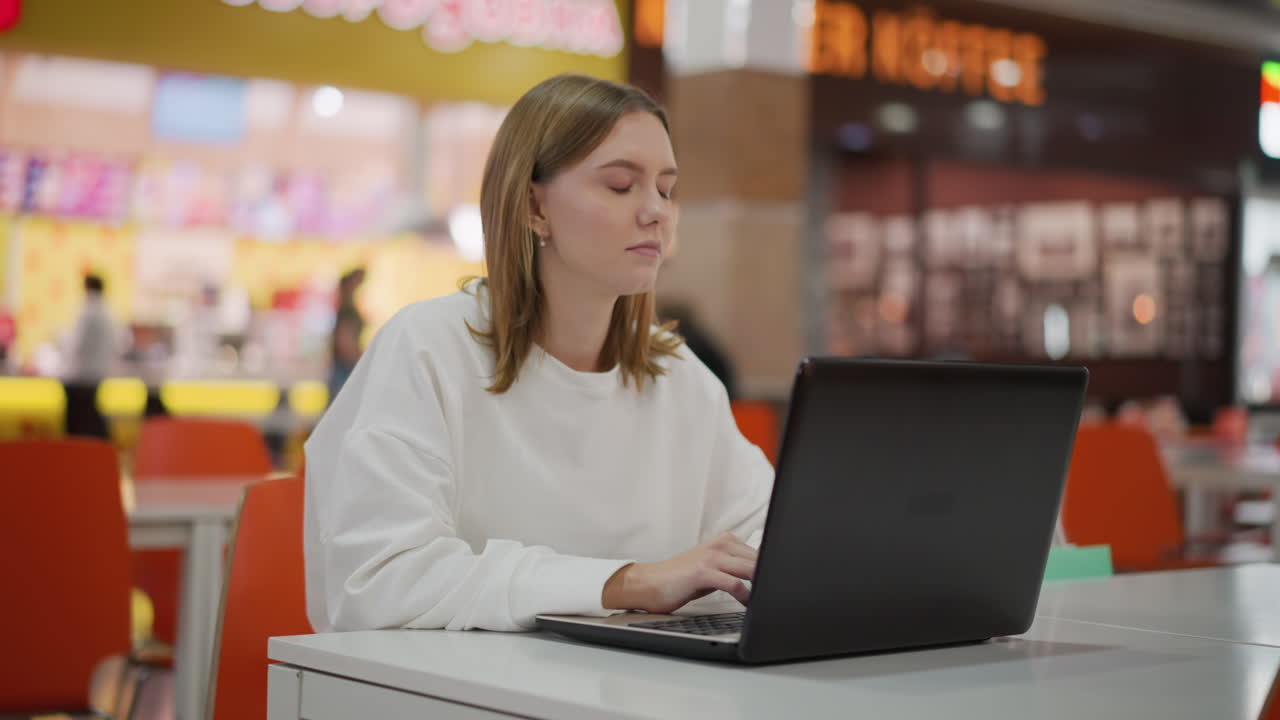trabajador remoto escribiendo en una computadora portátil en el patio de comidas del centro comercial, sentado en una mesa con un fondo interior moderno con colores vibrantes, señalización de marca y actividad borrosa
