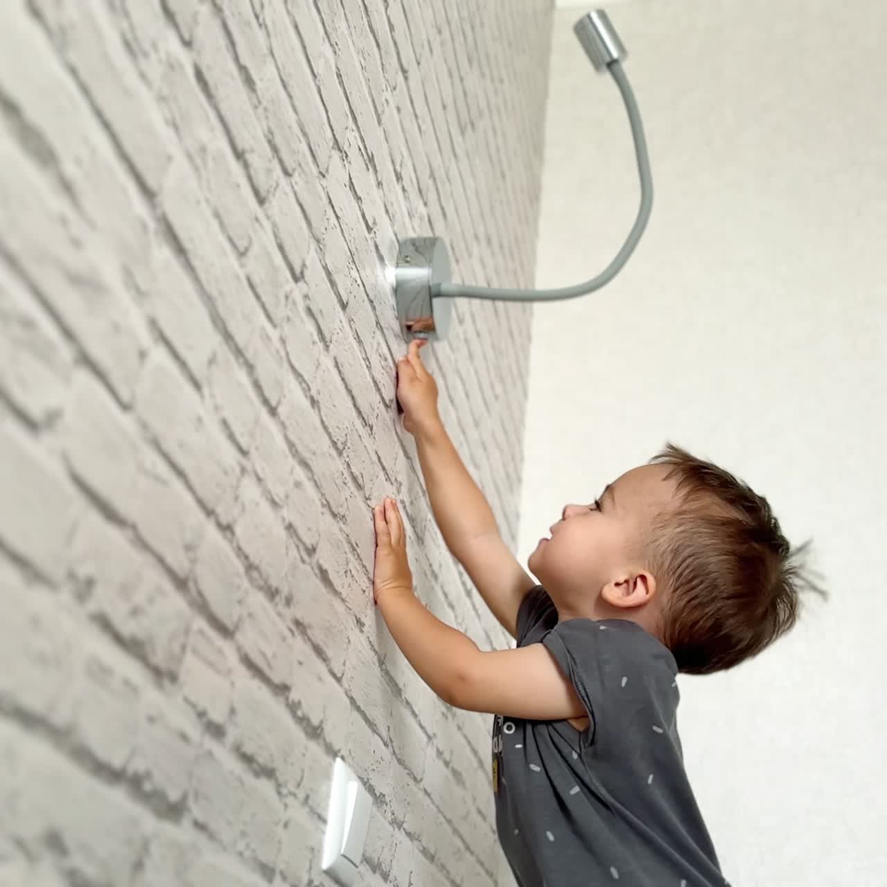 Energetic baby boy wearing grey t-shirt reaching the lamp on the wall. Happy kid pushes the button on the lamp. Low angle view