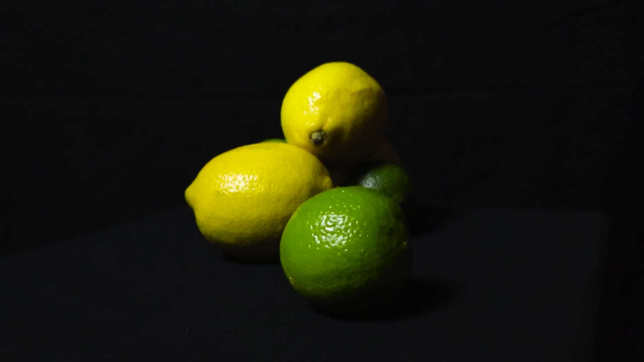 Gorgeous footage of yellow lemons and green vibrant colored lime citrus fruit on a dark black studio background while footage spins around them slowly. The fruit are under good studio lights.