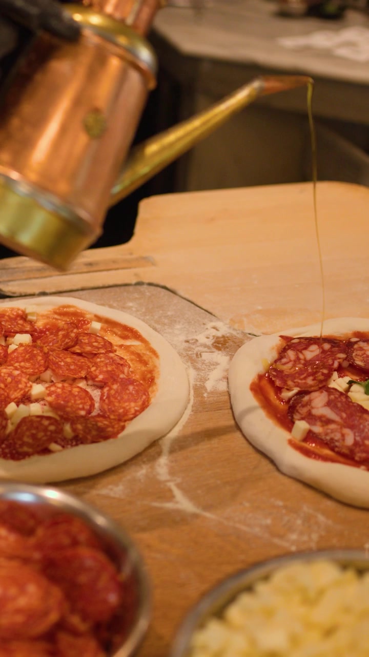Uno chef con guanti neri aggiunge salame piccante e olio a pizze crude su un bancone di legno.