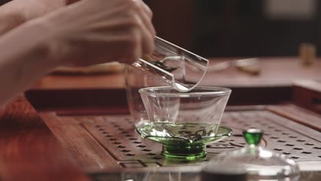 mujer asiática pone hojas de té verde de una bandeja de vidrio en un tazón de gaiwan chino en una mesa de té de madera tradicional