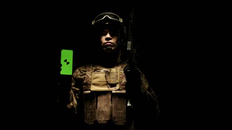 military soldier man in a helmet, army uniform and with rifle shows a green phone screen with the app, standing in the dark studio