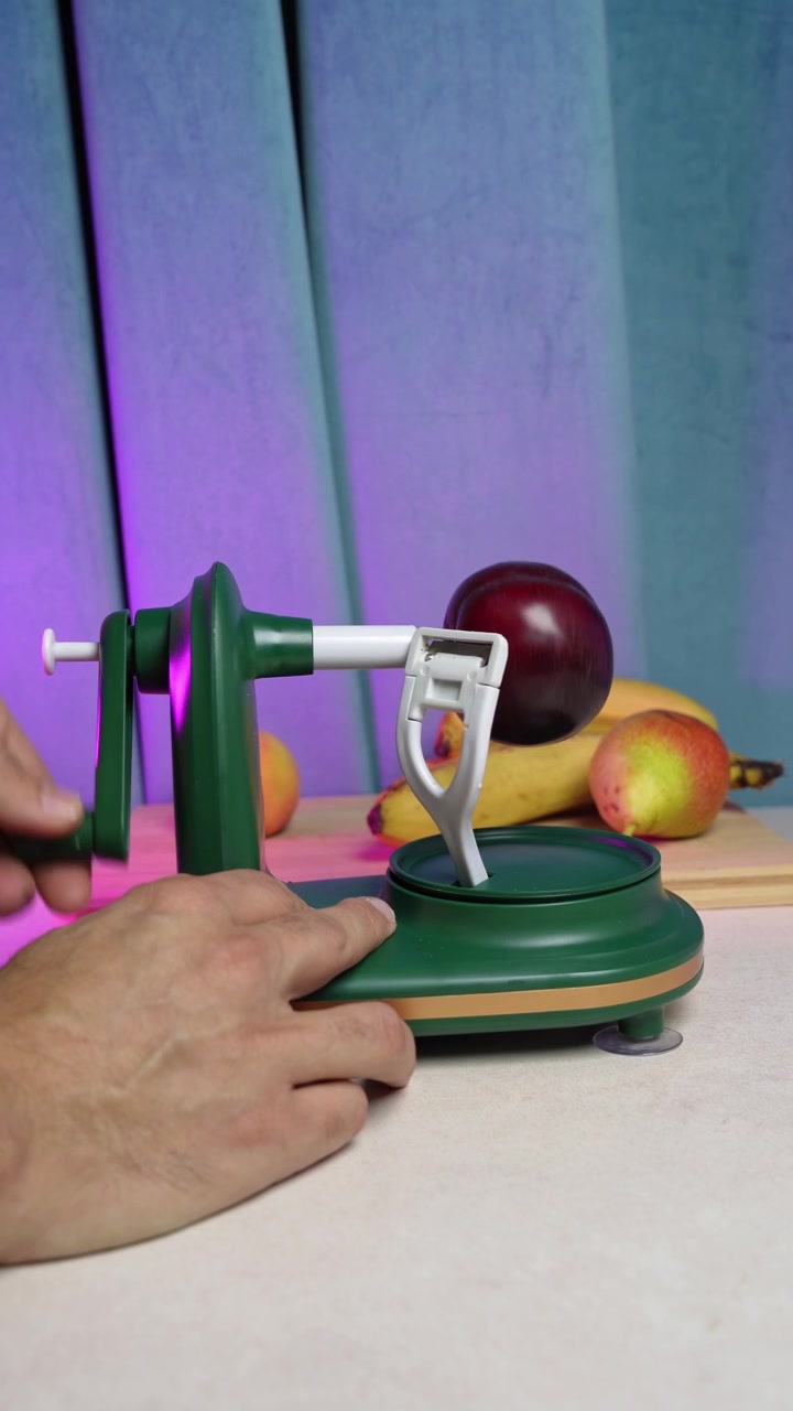 Scatto verticale ravvicinato di una persona che usa un pelapatate rotante manuale verde per sbucciare un frutto rosso fresco. Il pratico gadget da cucina è su un bancone sotto un'illuminazione da studio colorata, con banane sullo sfondo.