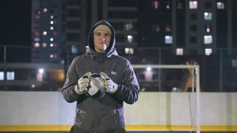 young boy in hoodie with gloves holding soccer ball and showing a subtle smile on urban sports field under night lights, background includes high-rise buildings, goalpost, and bokeh light effects