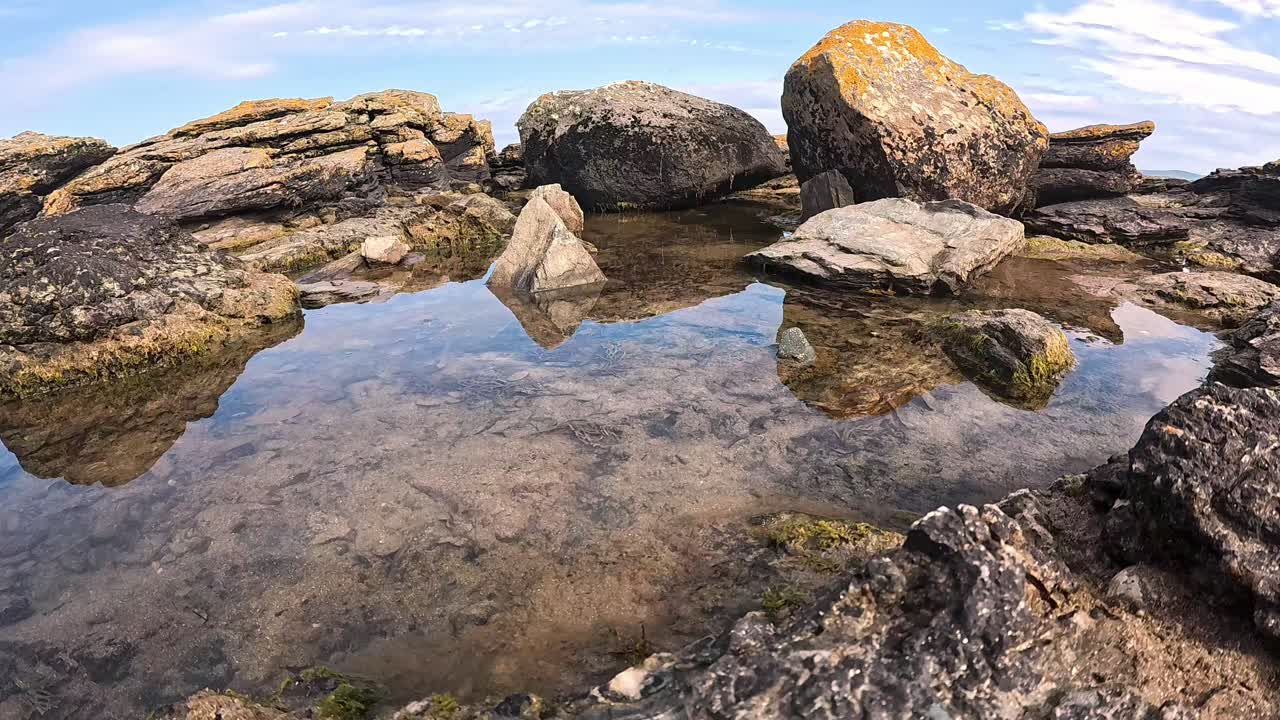 Una telecamera scorrevole a sinistra rivela la costa rocciosa di Penrhos, incorniciata da cieli soleggiati che si estendono oltre le pozze di marea, con l'acqua specchiata come soggetto principale della scena. Ideale per la narrazione naturalistica o atmosferica.