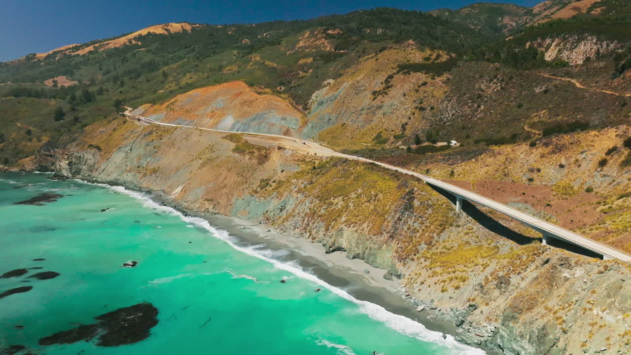 Amazing turquoise water of the Pacific with foamy waves arriving to the rocky shore. Mountains at the coastline with highway and bridge on scorching summer day. Top view.