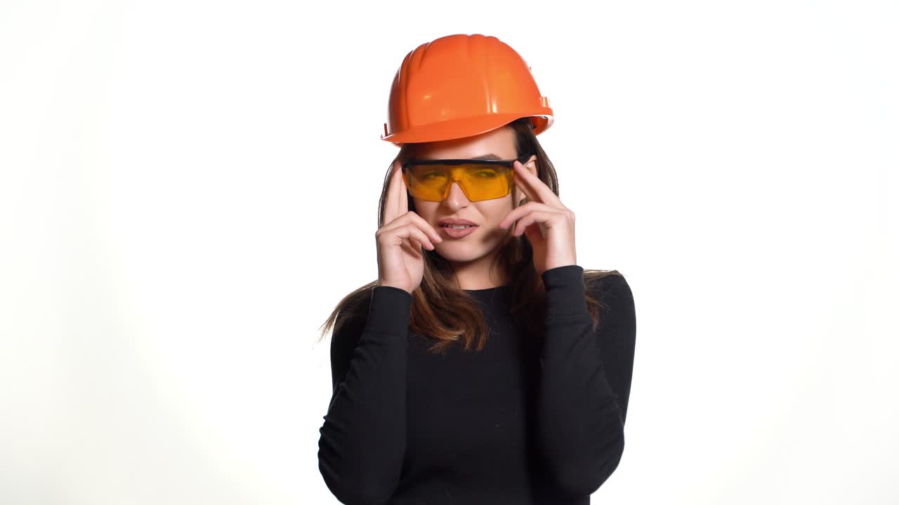 A joyful woman in orange helmet for building is trying on goggles and posing in it on a white background in the studio.