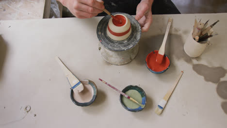 overhead view of male potter using brush to paint pot on potters wheel at pottery studio