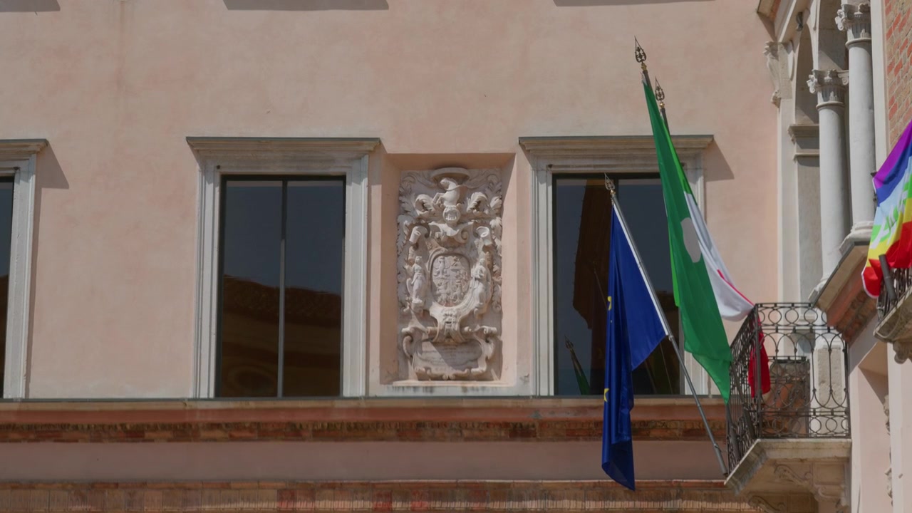 Stemma scolpito nella pietra tra due grandi finestre sulla facciata di un edificio. Primo piano delle bandiere dell'Unione Europea, della regione Lombardia e della pace arcobaleno che pendono dal balcone di un palazzo storico su un