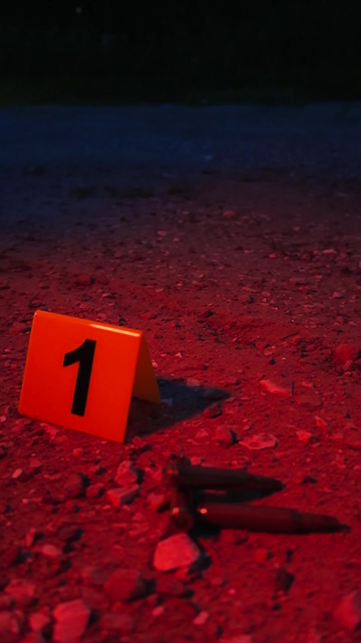 Indagine sulla scena del crimine di notte con marcatori di prove accanto a bossoli di proiettile su terreno fangoso, illuminata da luci rosse e blu lampeggianti di un'auto della polizia durante un intervento di emergenza.