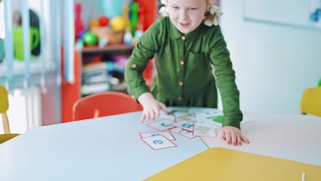 ein glückliches kleines mädchen an einem tisch. ein neugieriges kind, das allein im klassenzimmer sitzt und alle karten auf dem tisch zu sich nimmt. ein süßes vorschulkind mit bildern im innenraum.