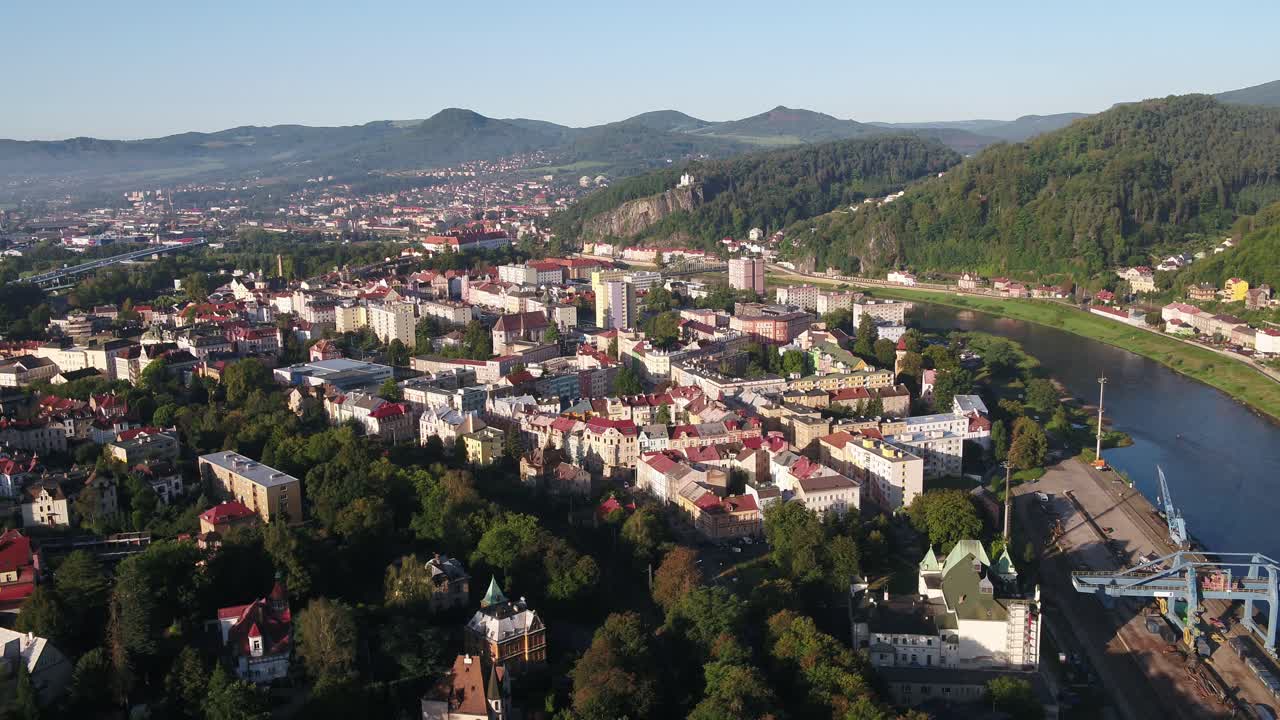 Drone zooms out revealing colorful buildings in Decin city center, surrounded by forested hills and clear skies