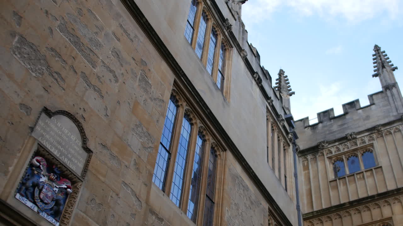 Facciata di un edificio all'Università di Oxford con alte finestre gotiche e muri in pietra consumati, ripresa da un'angolazione bassa che enfatizza le linee verticali e i dettagli storici del college.