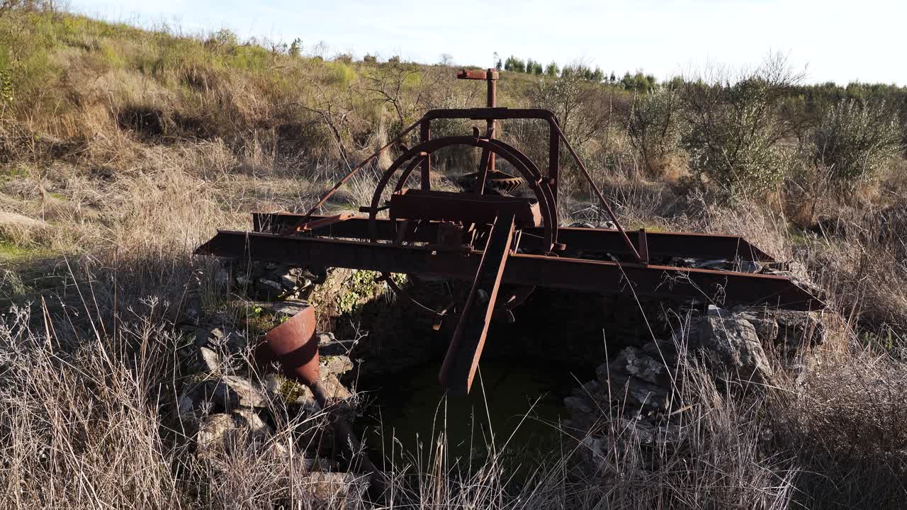 Primo piano di un vecchio motore manuale coperto di ruggine, situato accanto al pozzo di una fattoria, con ingranaggi e tubi usurati un tempo utilizzati per estrarre l'acqua per l'irrigazione, che mostrano anni di utilizzo e storia agricola