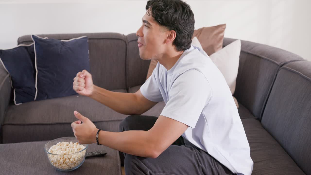 Asiatisk mand sidder på grå sofa, reagerer på skærmen, klapper mod popcorn og fjernbetjening, med plads til tekst.