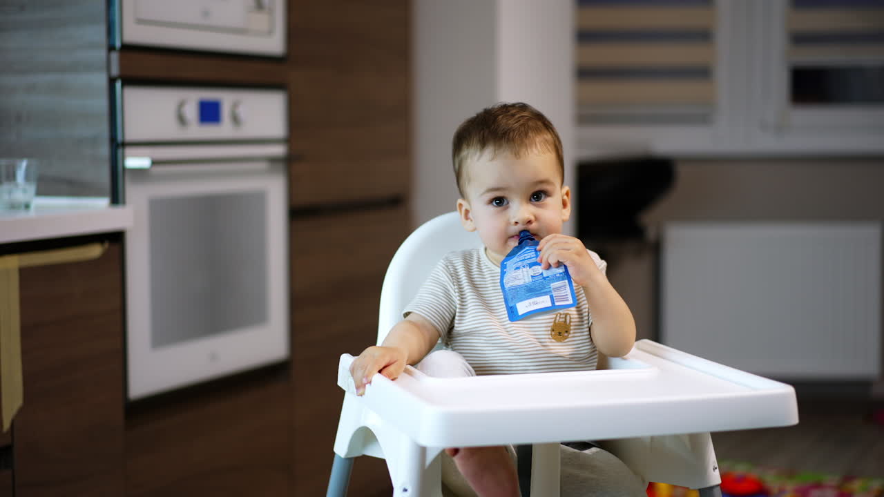 Cute Caucasian baby boy sits in a high chair. Toddler is holding a fruit puree pack in his mouth and smiles adorably to the camera. Blurred backdrop.