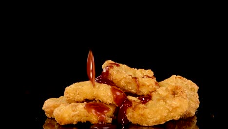 golden, crumbed chicken pieces are drizzled with dark sauce in slow motion on a glossy black surface, under dramatic studio lighting with a static camera