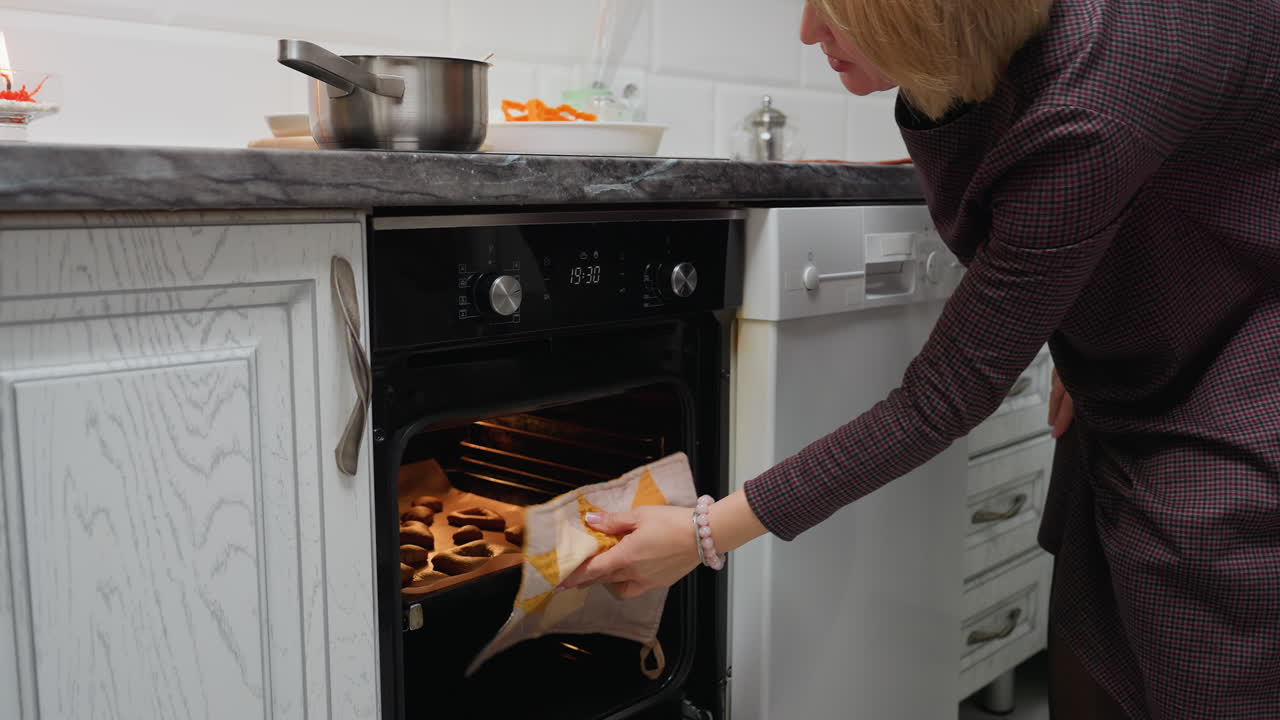 Signora che usa uno strofinaccio per togliere i biscotti cotti dal forno con una pentola sul piano di lavoro, biscotti su carta da forno, cucina ben illuminata, biscotti caldi e dorati appena sfornati