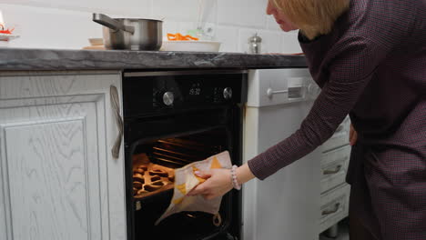 lady using rag to remove baked cookies from oven with pot on countertop, cookies placed on parchment paper, well-lit kitchen, warm and golden cookies freshly baked