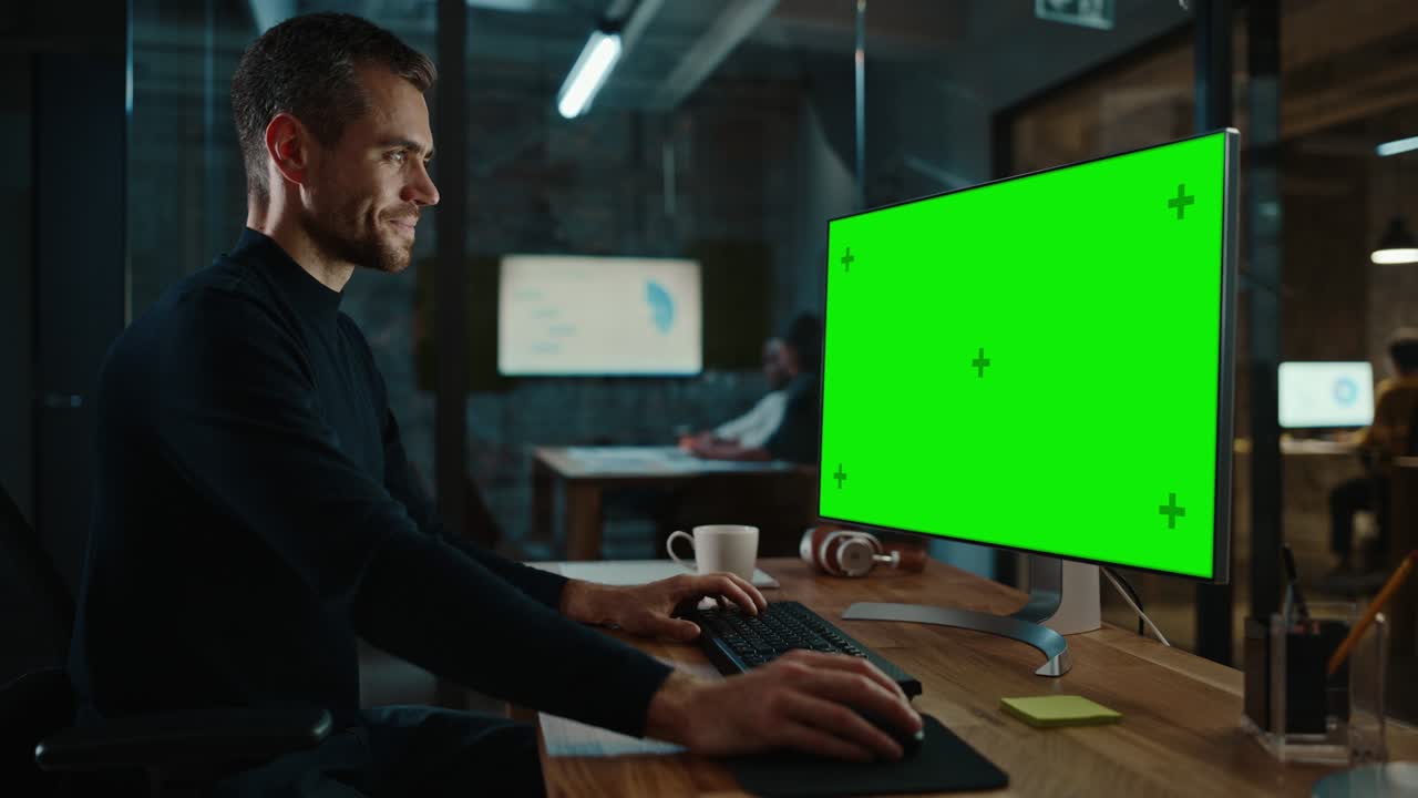 Young Handsome Specialist Working on Desktop Computer with Green Screen Mock Up Display in a Busy Creative Office with Colleagues. Male Manager with Trimmed Beard is Wearing a Casual Black Jumper.