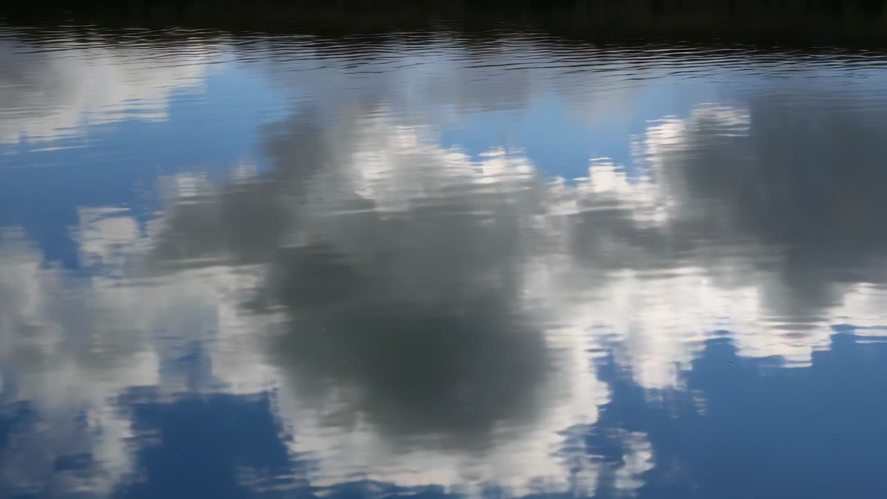 Pantulan awan yang beriak berkilauan saat angin sepoi-sepoi memicu riak horizontal kecil di seberang danau