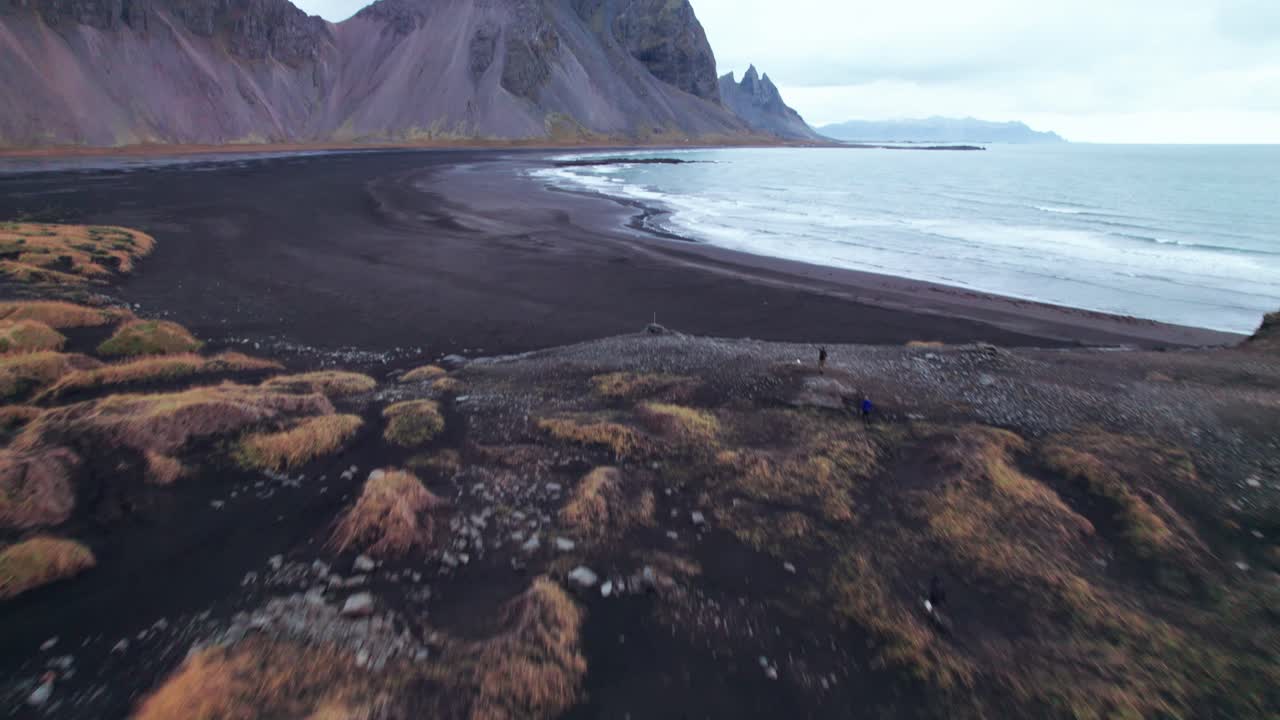 aerial: tilt up revela tussocks, playa de arena negra, y vestrahorn, con sus icónicos picos dentados y altos contrafuertes, se eleva dramáticamente desde esta oscura extensión como una colosal catedral de piedra