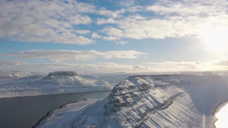 incredible wide bird's aerial drone view of iceland snowed westfjords on sunny bright day, mountain range landscape surrounding large calm icelandic ocean river water, circle pan