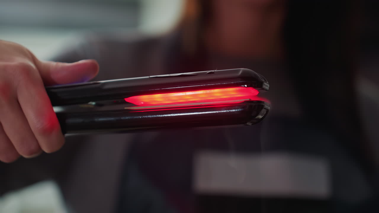 Primer plano de estilista caucásico sosteniendo una plancha brillante, placas LED rojas visibles, delantal de peluquería al fondo, aparato moderno comprobado para temperatura y seguridad, agarre y control precisos