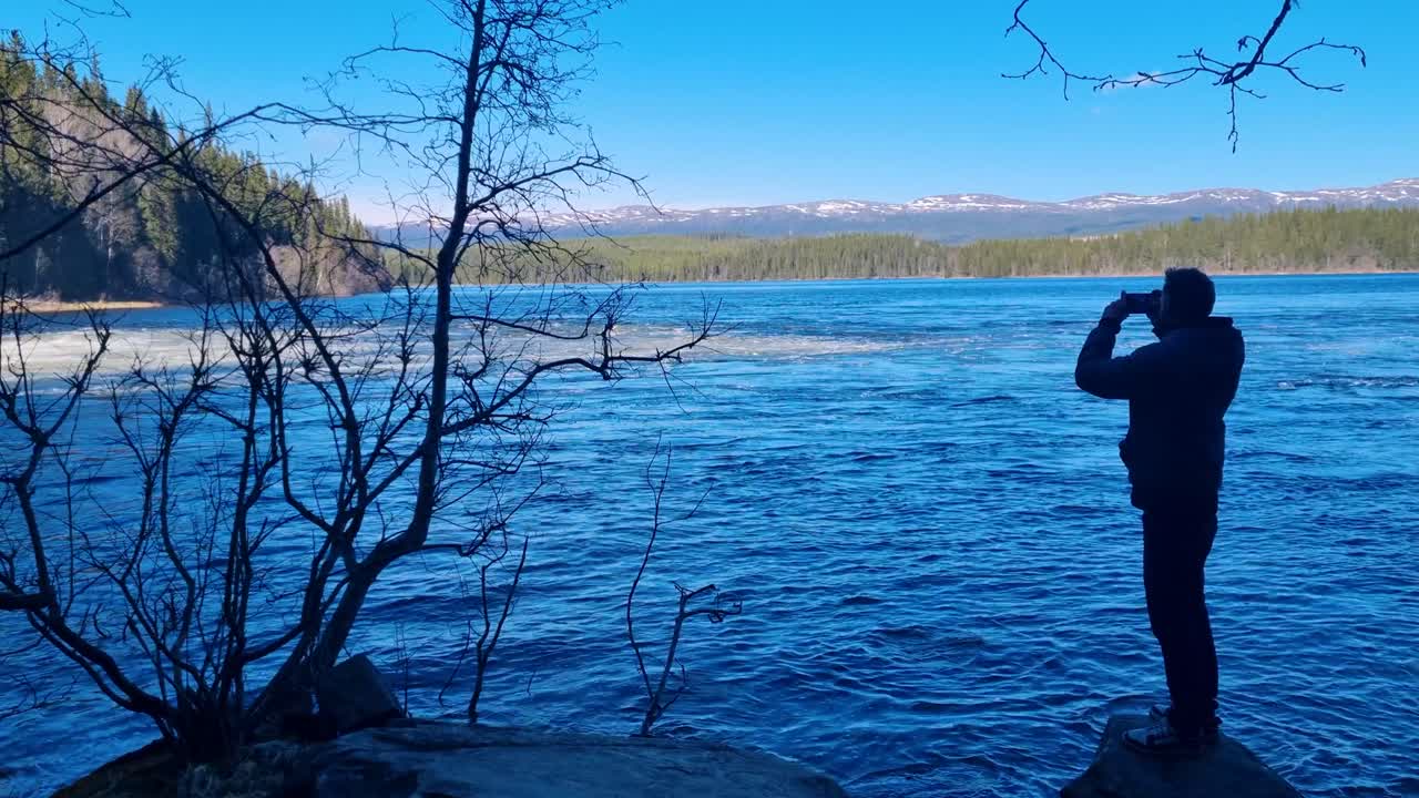 A man stands in the shade on a rock, nearly silhouetted, capturing where rushing water meets a deep blue lake, set in a picturesque Scandinavian landscape on a clear, sunny day