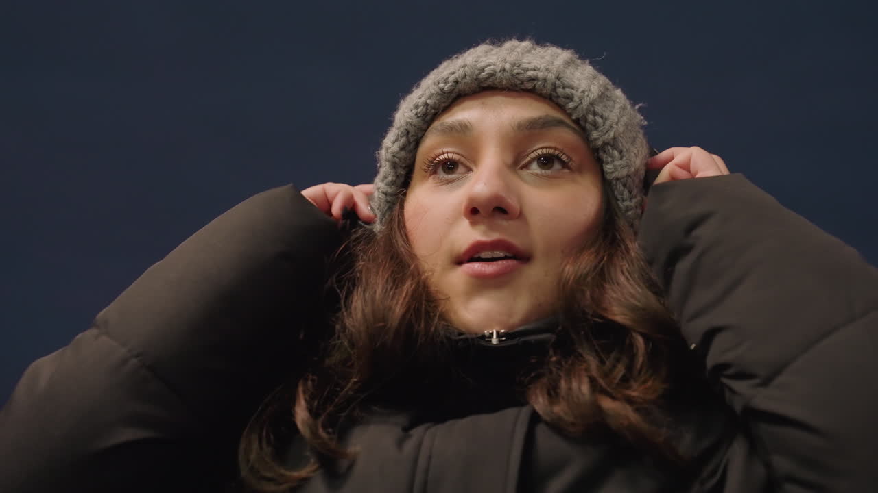 Closeup riverside urban woman with headphones syncing to beat, hands adjusting earcups, eyes brightening, subtle body sway, city lights and bridge in background, energetic cozy night vibe