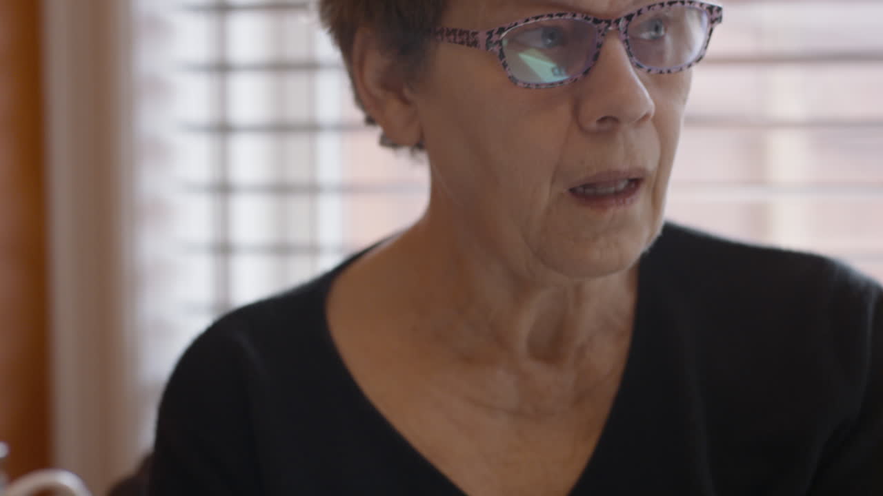 An elderly woman's face focused on gently self-massaging the spasticity in her hemiplegic right hand. She looks up from her hands, giving a sigh of frustration, or impatience.