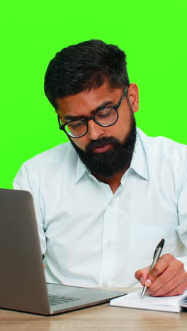 indian man writing ideas in notebook while working on laptop focused multitasking planning concept