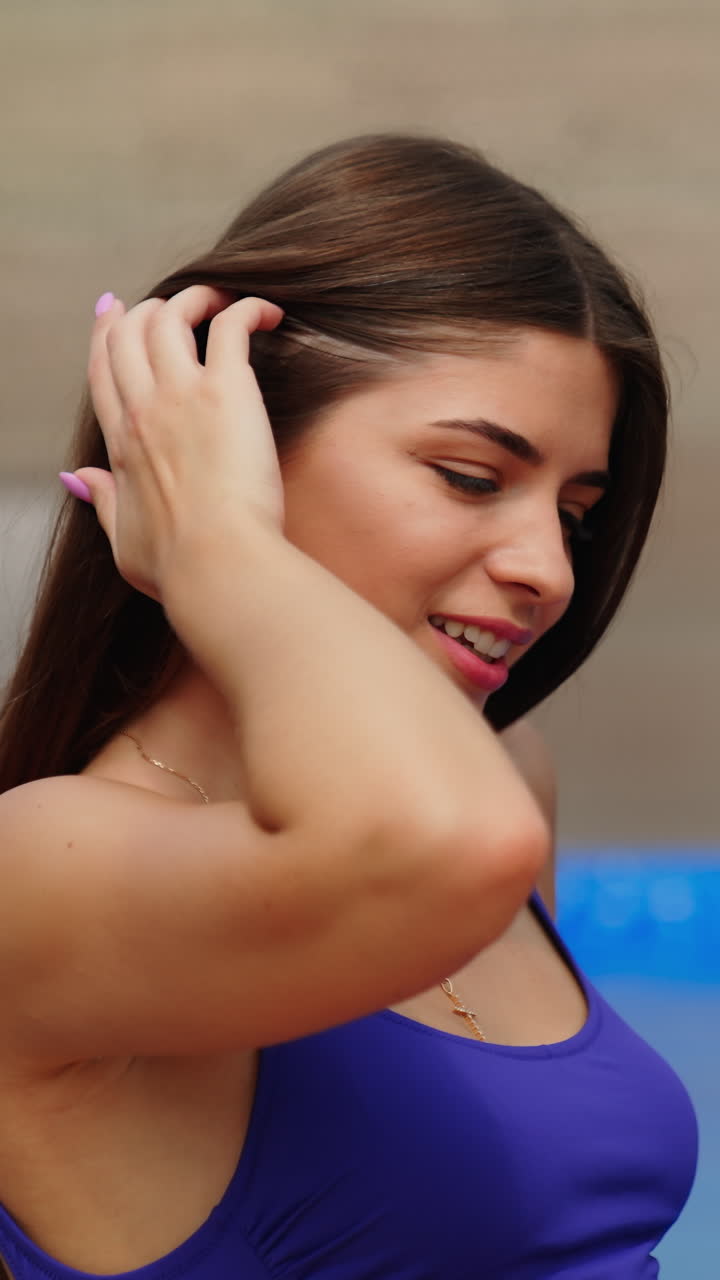 Beautiful brunette girl with long hair sitting at the pool. Young woman in purple swimsuit posing to the camera. Vertical video